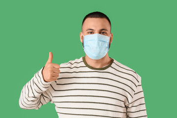 Young man in medical mask showing thumb-up gesture on green background