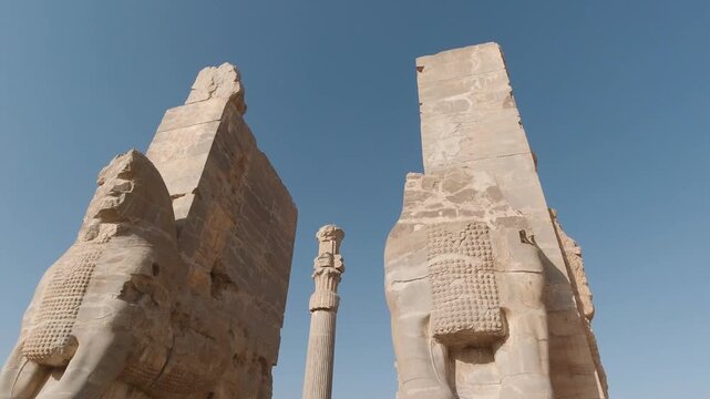 Low-angle tracking shot between the colossal guardian bulls of the Gate of All Nations at Persepolis, Iran, revealing fluted columns and Achaemenid stonework under a clear blue sky.