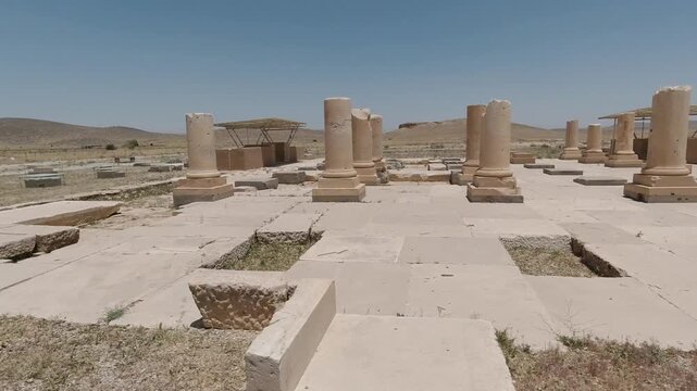 Wide view of Pasargadae&rsquo;s colonnaded hall: stone columns and bases aligned across the palace floor, excavated grid visible in bright summer light with arid hills on the horizon.