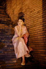 a young woman in traditional thai dress sitting on brick stairs at night during yi peng and loy krathong festival, smiling softly under warm lantern light that glows against the ancient brick wall