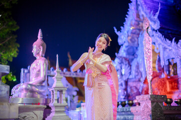 young woman wearing a traditional thai dress smiling and greeting with wai gesture in front of wat sri suphan temple during yi peng and loy krathong festival night