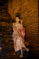 a young woman in traditional thai dress sitting on brick stairs at night during yi peng and loy krathong festival, smiling softly under warm lantern light that glows against the ancient brick wall