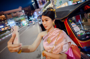 a young woman wearing traditional thai dress taking a selfie beside a colorful tuk tuk on a lively street at night during yi peng and loy krathong festival in chiang mai