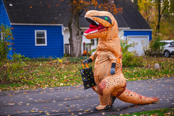 Person in inflatable dinosaur costume walking along suburban street with a Halloween candy bag. © StockMediaSeller