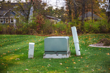 Utility boxes on a residential lawn surrounded by autumn foliage and houses in the background during fall season.