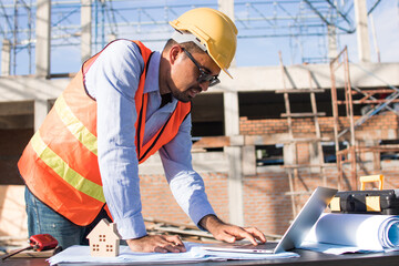 Construction worker wearing a working suit with helmet, holding a pen and looking at the papers with architectural site blueprint placed on the table.