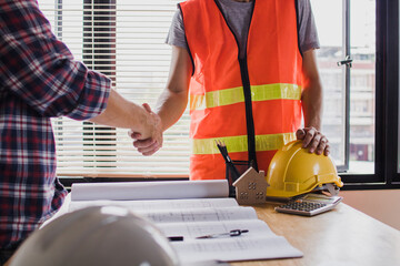 Two construction professionals shaking hands over blueprints and safety equipment signifying a successful project agreement and partnership