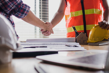Two construction professionals shaking hands over blueprints and safety equipment signifying a successful project agreement and partnership