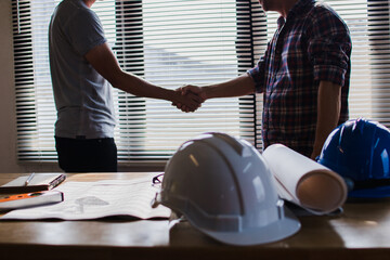 Two construction professionals shaking hands over blueprints and safety equipment signifying a successful project agreement and partnership