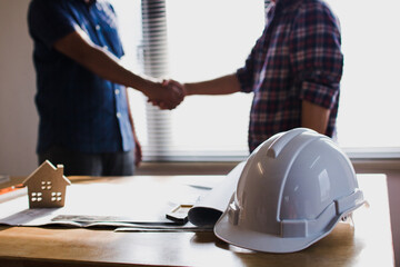 Two construction professionals shaking hands over blueprints and safety equipment signifying a successful project agreement and partnership