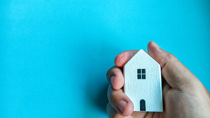 Minimalist concept image showing a hand gently holding a small wooden house on a blue background, symbolizing home ownership, safety, real estate, family care, and housing security.