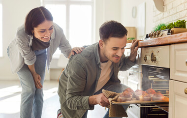 Happy smiling young couple baking together in kitchen, enjoying fresh homemade muffins just out of oven. Cheerful man taking tray of golden cupcakes out of oven while woman watching with excitement.