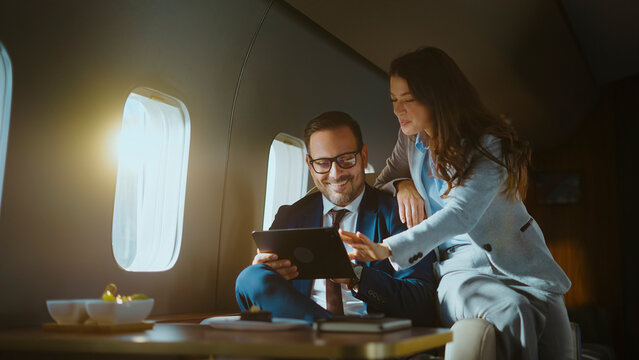 Business executive male and female colleagues collaborating and analyzing data on a digital tablet while flying in a luxurious private airplane cabin, representing successful corporate travel - Powered by Adobe