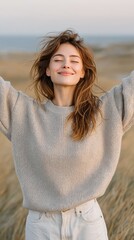 Young Woman With Curly Hair Wearing A Textured Sweater And White Pants With Arms Raised In A Grassy Field With Ocean In The Background During Golden Hour