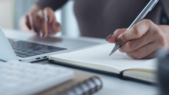 Close up of business woman working on laptop, searching the information and writing on notebook, business planning. Student studying online class via laptop, e-learning, online research