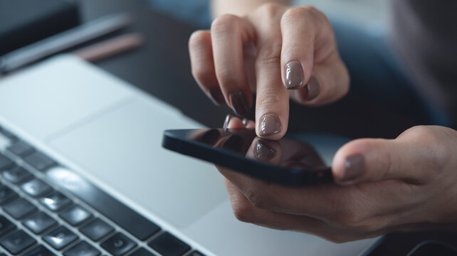 Close-up iof casual business woman using smartphone while working on laptop computer at home office, working remotely, freelance at work, online job, brown tone