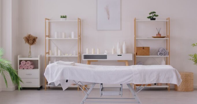 Bright empty massage room with white massage table and wooden shelves with beauty skincare products. Modern interior of spa center, beauty salon, aesthetic clinic of dermatology and cosmetology.