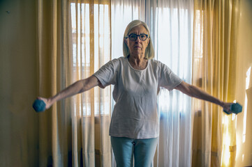 Elderly Woman Doing Arm Exercises With Dumbbells At Home In Morning Light By Window Curtains