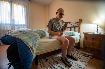 Senior Man Relaxing in Bedside Bedroom Using Smartphone in Cozy Home Setting During Morning Light