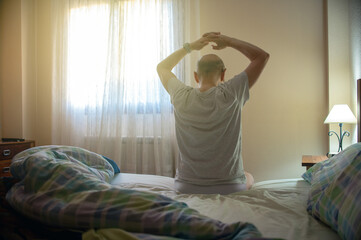 Man Sitting On Bed Stretching In Bedroom With Hands Raised At Morning Light