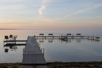 Empty dock with benches over calm lake during soft pastel sunrise.