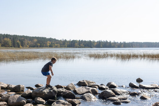 Boy balancing on rocks at calm lakeshore under clear sky.