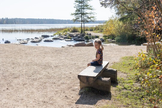 Smiling child sits on wooden bench near lakeshore on sunny day.