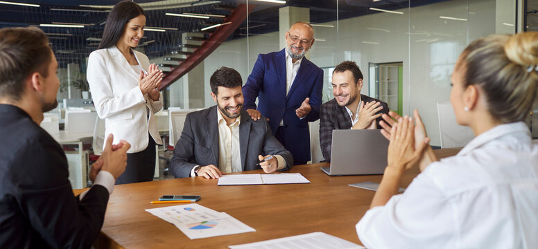 Applause from business team for success signing of contract on meeting in office. People clapping hands, sitting and standing at table, happy manager with pen looking on agreement document - Powered by Adobe