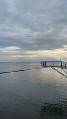 Fototapeta premium Serene Seascape with Structure: An expanse of calm water mirrors the overcast sky, with a modern structure poised on the horizon, hinting at a blend of nature and human design.