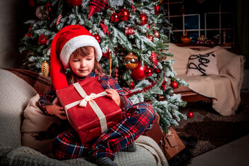 Happy little girl child in pajamas and festive santa hat smiling while sitting near christmas tree with gift box. Magic. New Year's eve. St. Nicholas Day. Present. Time of holidays and miracles.	