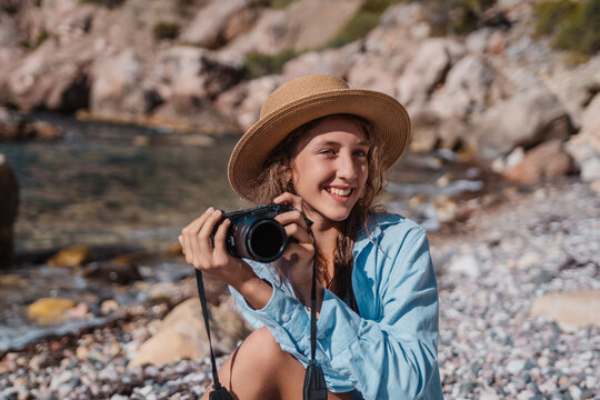 Girl, photographer, beach: Young woman smiling while holding a camera on a rocky coastline vacation.