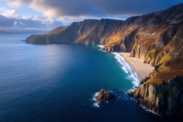 Coastal cliffs meet a sandy beach bordered by a dark blue sea under a partly cloudy sky