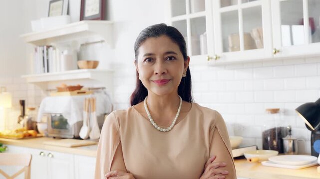 Smiling confident stylish Asian mature senior aged woman standing at home kitchen. Looking at camera arms crossed