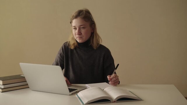 Successful young woman freelancer with cerebral palsy studying and working, writing notes in notebook while using laptop and books, integrating disability into productive work-study environment.