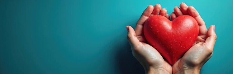 Fototapeta premium Two hands cradle a bright red heart. This symbol of love and care rests gently against a calm teal backdrop. Its smooth surface is dotted with tiny water droplets.