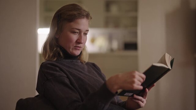 Determined woman with cerebral palsy and spasticity reading book, carefully flipping through pages in cozy evening home setting, emphasizing importance of learning and resilience, slow motion.