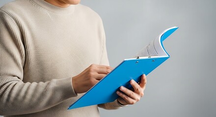 Close up of a person wearing a textured sweater holding and reading a bright blue folder or binder with a curved page on a neutral background