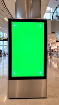 Blank digital advertising display with a chroma key green screen and tracking markers inside a bustling airport hall with escalators