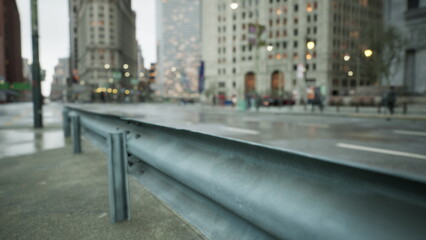 Rain falls gently on a bustling city street, creating reflections in puddles. Blurry figures walk by, while tall buildings loom in the background, adding to the urban atmosphere.