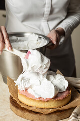 A chef spreads fluffy meringue over a pink-filled tart base using a spatula. The dessert sits on a wooden board lined with parchment paper. A close-up of the cake decorating process, highlighting crea