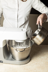 A person in a white chef’s shirt pours liquid from a stainless steel pot into a planetary mixer bowl with whipped ingredients. Close-up of the baking/cooking process on a clean kitchen countertop.