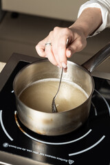 A person stirs a bubbling liquid in a stainless steel saucepan on an induction cooktop. Close-up of the cooking process, focusing on the hand and spoon action. The liquid appears to be simmering.