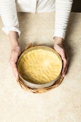A pair of hands holds a round baking pan with shortcrust pastry base, lined with parchment paper. Ready for baking, set on a light marble countertop. Clean, minimalist kitchen scene, close-up shot.
