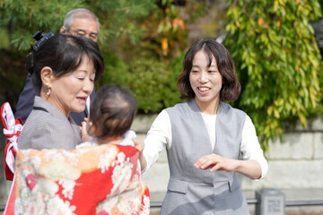 A woman is holding a baby and smiling at another woman. The two women are standing in front of a green bush