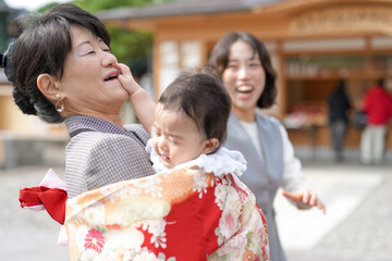 A woman is holding a baby in a red and white blanket. The baby is crying, and the woman is trying to comfort it. Another woman is standing nearby, smiling at the scene
