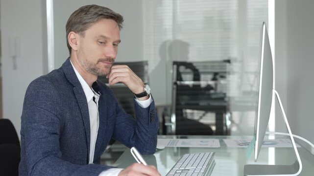 Businessman in a blue jacket thinking and brainstorming ideas while working on computer and taking notes in modern office. Business people concept