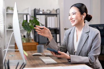A cheerful young Asian businesswoman in a grey suit on a video call, wearing an earpiece and gesturing confidently while communicating at her modern office desk.