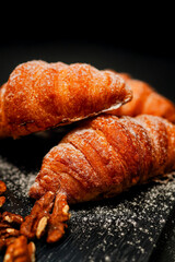 A vertical, moody close-up of golden, flaky croissants, generously dusted with powdered sugar, stacked on a dark board with scattered walnuts, perfect for bakery promotion.
