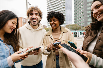 Happy young group of diverse friends having fun using mobile phones at city street, texting...