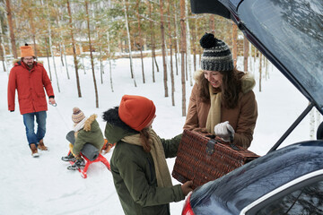 Caucasian young woman and Caucasian child unloading picnic basket from car trunk while smiling, with Caucasian man pulling Caucasian child on sled in snowy forest background